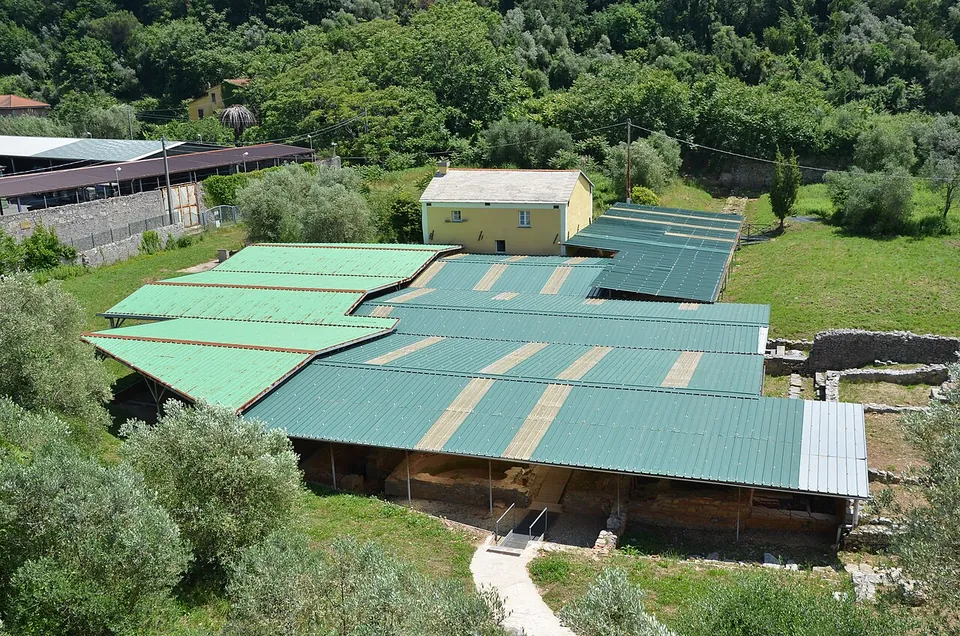 Seen from above, a group of long green metal roofs covers the excavations in a grassy hollow, with a yellow two-storey house behind them, olive trees in the foreground and a wooded hillside beyond.