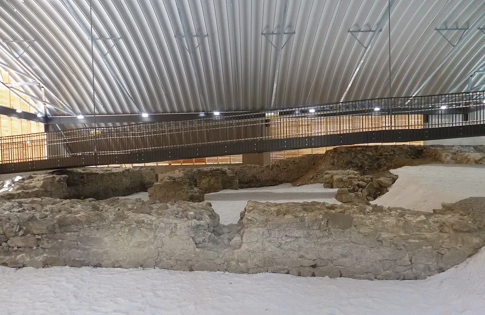 Low walls of rough stone laid out on pale gravel inside a large modern hall with a curved corrugated metal roof, crossed by a raised steel walkway with railings.