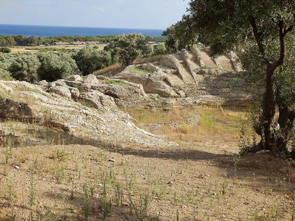 Weathered banks of pale stone curving round a bare earth floor, with olive trees at the edge and the sea visible in the distance.