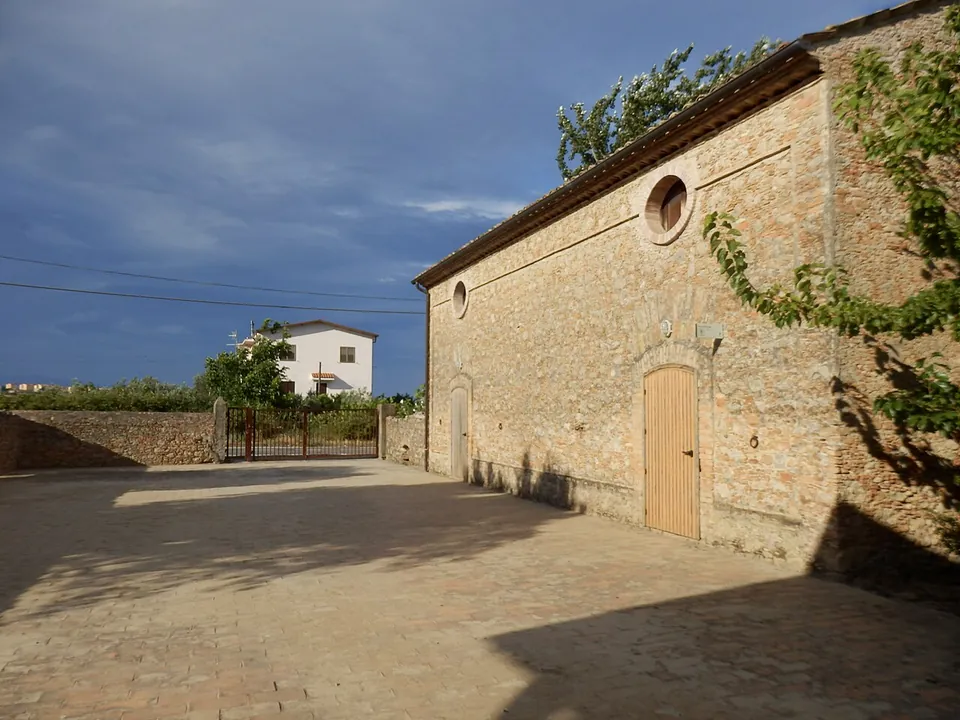 A paved courtyard beside a long stone building with a round window and an arched wooden door, with an iron gate in a low wall and a white house beyond.