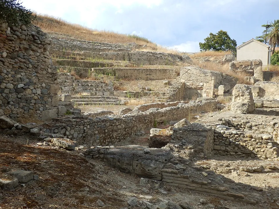 Rows of stone seating rising up a grassy slope above the excavated stage area, with walls of rough rubble stone in the foreground and a small white house on the hilltop behind.