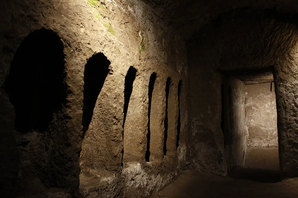 A rough rock wall lined with a row of tall, round-topped niches, lit from above, with a doorway at the end opening into a second chamber.