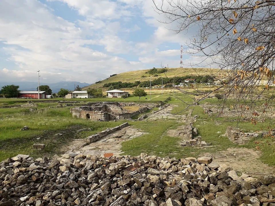 Excavated stone walls and strips of paving running across a grassy field, a heap of rubble in front, low white buildings and a hill with a mast behind.