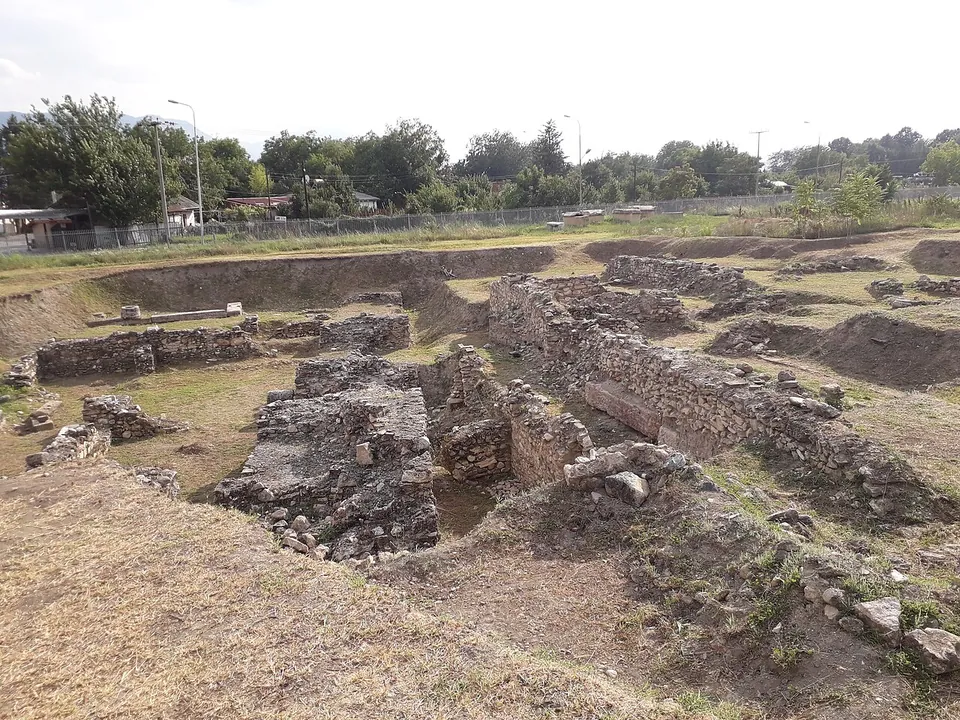 Rubble-stone walls of several rooms standing in a shallow excavation pit with grassy banks, a fence, street lamps and trees beyond.