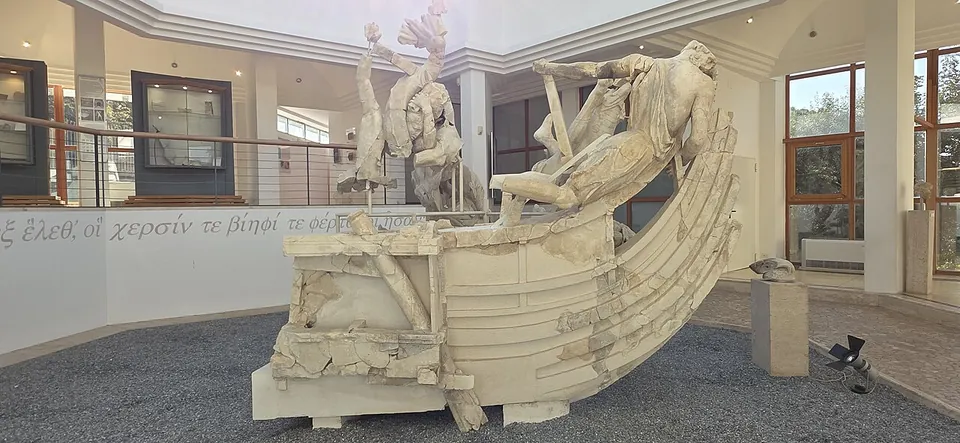 A large reassembled white marble sculpture in a museum hall, the stern of a ship with straining figures on its deck and a larger figure rising behind it, set on a bed of grey gravel, with a line of Greek lettering on the wall behind.