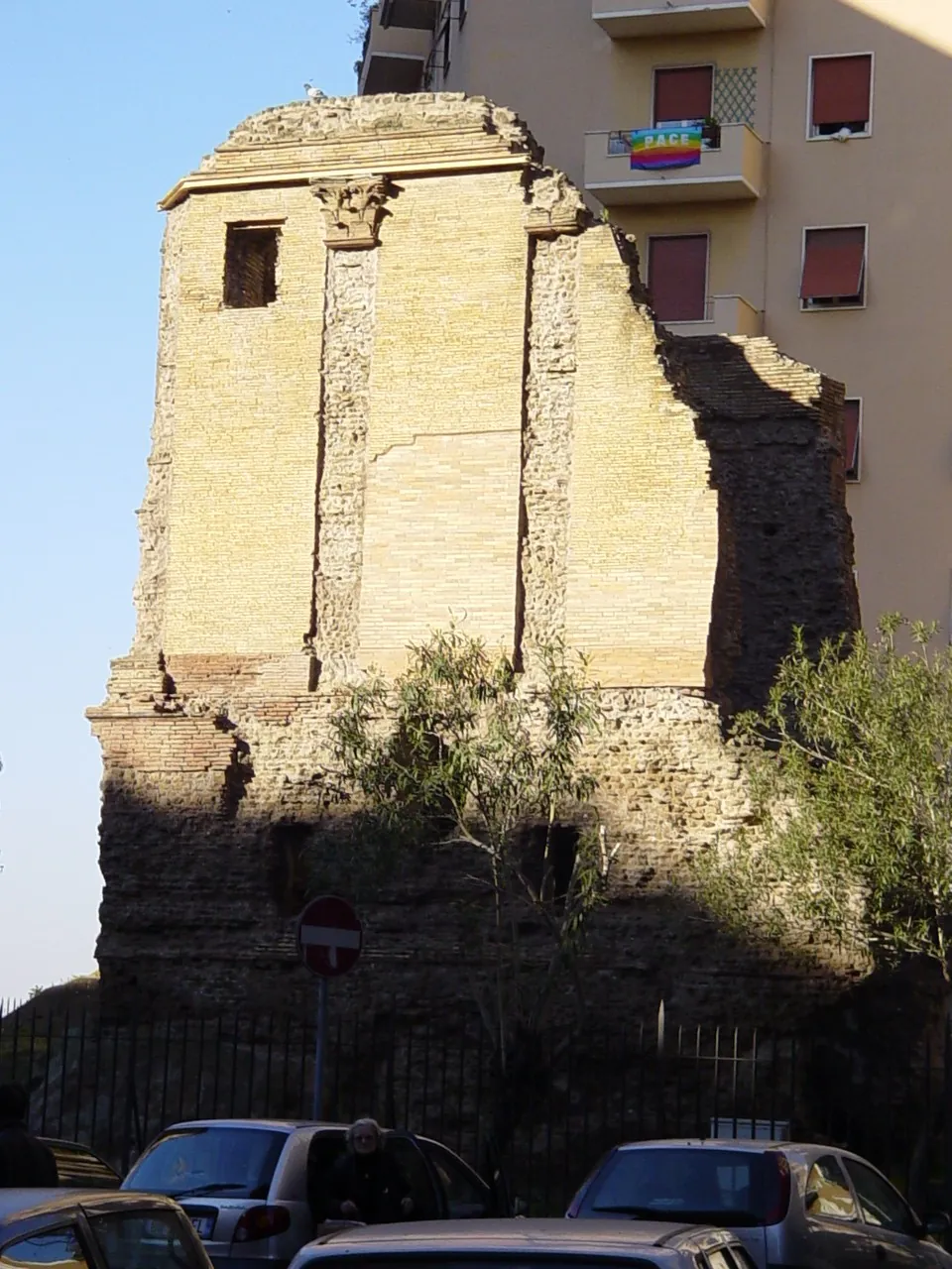 A tall ruined brick tomb with its front fallen away, pilasters and a small window on its side wall and a rough concrete core below, behind railings, with apartment blocks behind and parked cars in front.