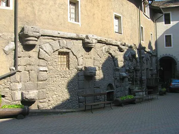 A row of large stone arches with carved stone brackets above them, built into the plastered wall of a later building, with a bench and plant pots in the courtyard in front.