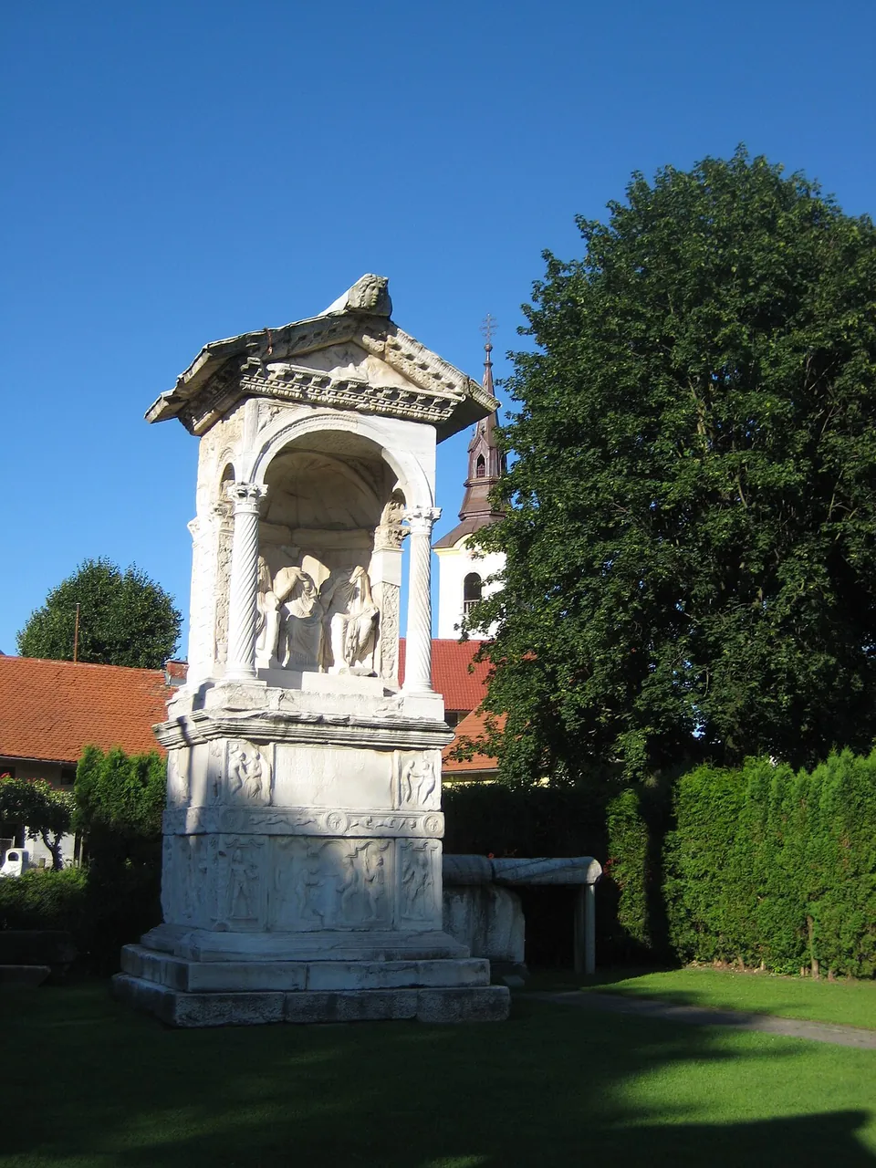 A tall white marble tomb against a blue sky, a pedimented roof on twisted columns sheltering carved figures, standing on a high base whose sides are covered in relief panels.