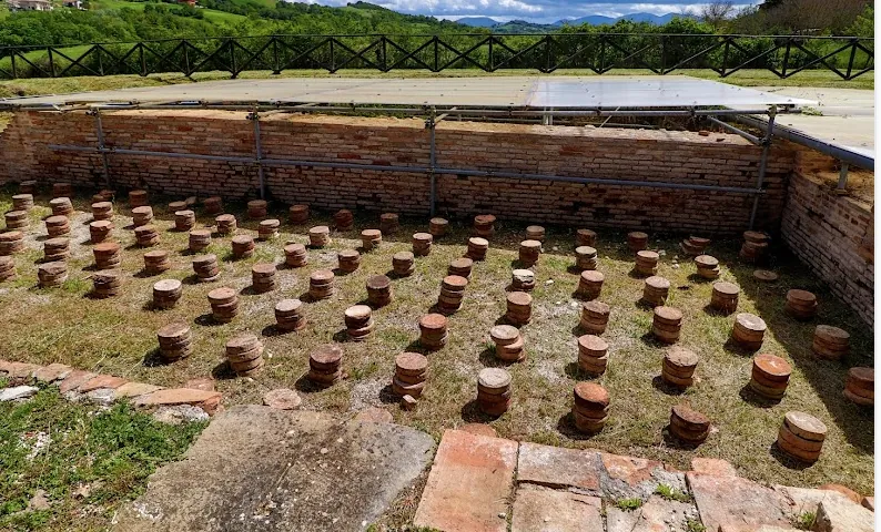 A sunken rectangular room with brick walls, its floor covered with rows of short round stacks of terracotta tiles, under a flat modern canopy and inside a wooden fence.