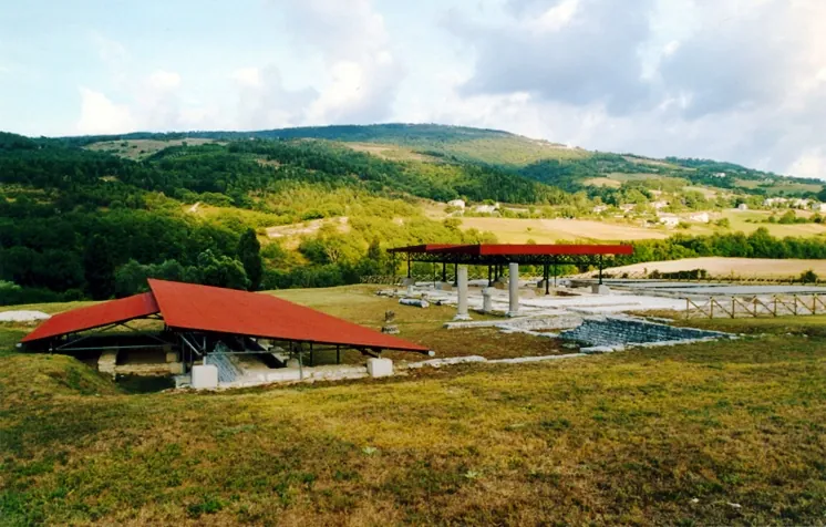 Excavated remains spread over a grassy terrace, two of them under modern shelters with red roofs on metal posts, a few standing columns among low walls, and green hills and farmland behind.
