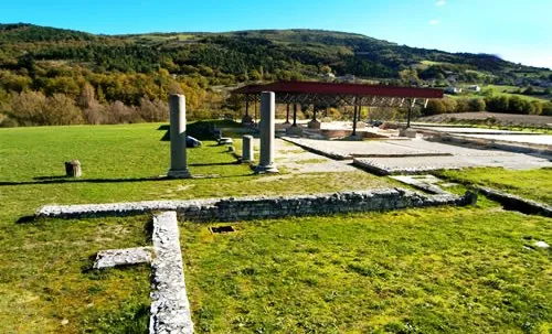 Two plain stone columns standing in mown grass beside the low foundations of walls, with a red-roofed shelter over excavated remains behind them and wooded hills beyond.