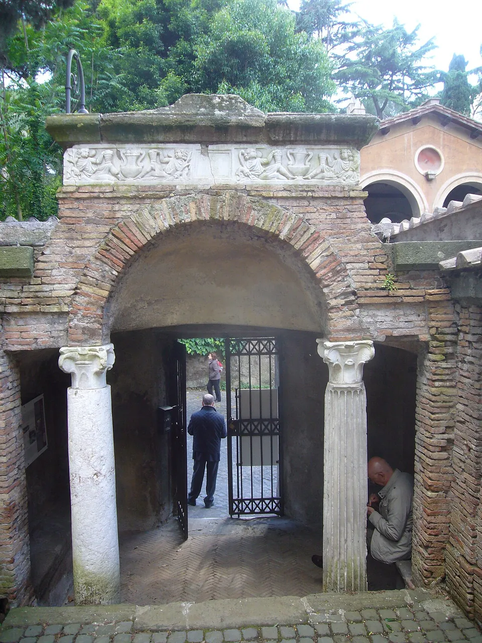 A small brick gateway with a round arch carried on two columns, a carved band above the arch, and an iron gate standing open onto the street beyond, where a man is walking.