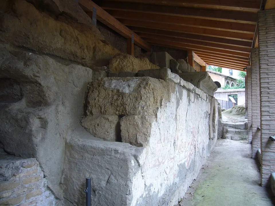 Large squared blocks of grey tuff stacked into a low wall against the rock, under a modern timber-and-brick shelter, with faint reddish marks on the plastered face and a narrow walkway running alongside.