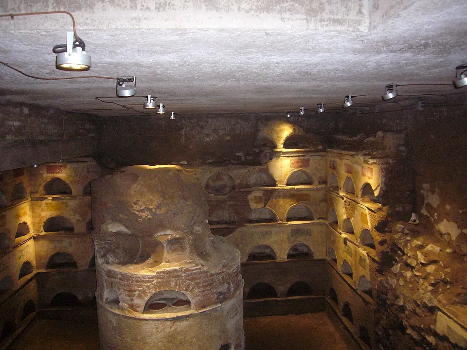 An underground room lit by spotlights, its walls lined with rows of small arched niches and a thick round pillar in the middle, itself pierced with niches.