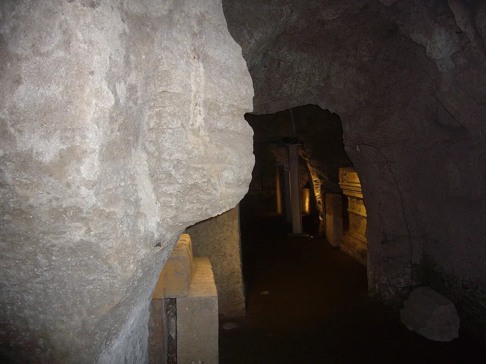 A dim gallery cut into pale grey rock, opening onto further passages lit by low lamps, with metal props standing against the walls and blocks of stone along the floor.