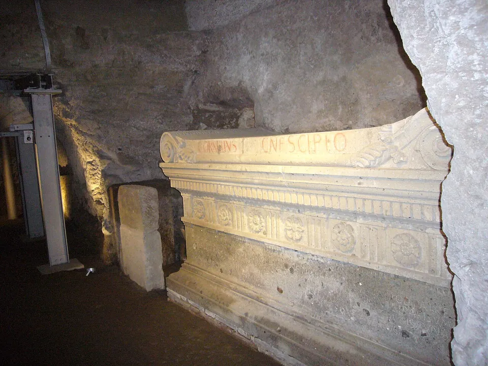 A large stone sarcophagus against a rock wall, lit from below, with scrolled ends to the lid, the words CORNELIVS CN F SCIPIO painted in red along it, and a frieze of rosettes between upright grooved panels.