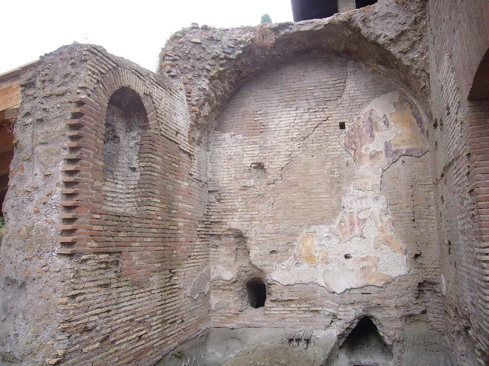 Tall ruined walls of brick and rubble, with a round-headed niche on the left and patches of painted plaster in faded colours on the wall at the back.