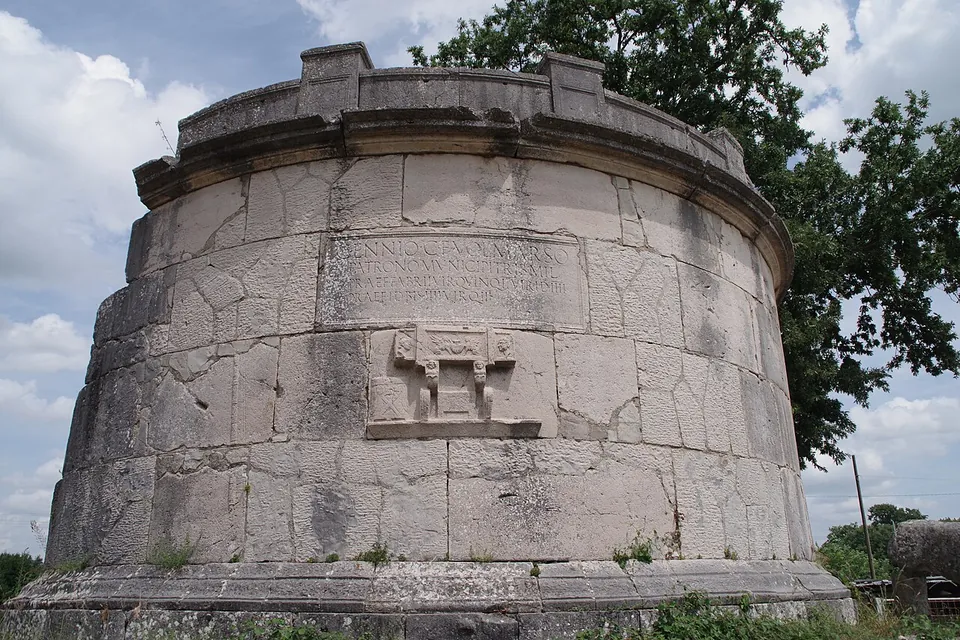 A squat cylindrical tomb of large pale stone blocks with a crenellated top, a long inscription panel across the drum and below it a carved relief of a folding seat between two objects, with trees behind.