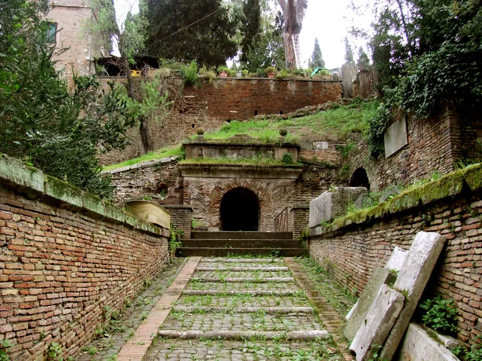 A sloping path paved with stone setts runs between two low brick walls to a few steps and a round-arched opening in a brick front set into a grassy bank, with further brick walls, trees and a building on the slope above.