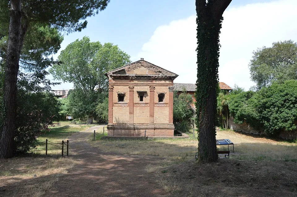 A square two-storey building of pale yellow brick with red brick pilasters and a low pitched roof under a pediment, standing in a grassy clearing among tall pines and trees, with an information board on the grass to its right.