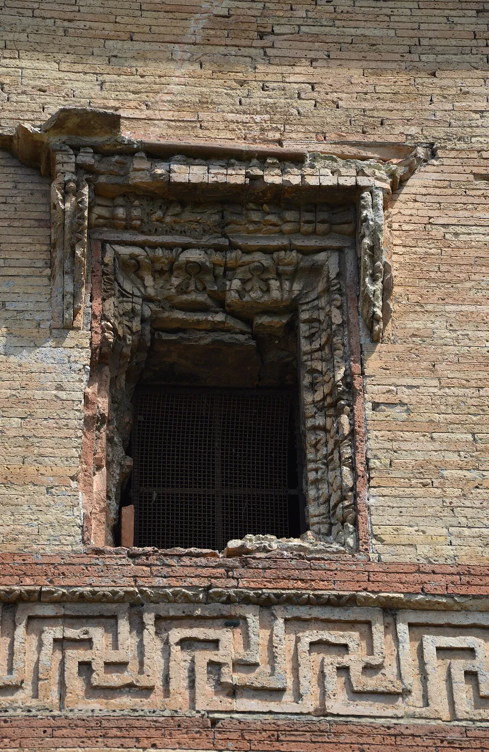 A small window in a wall of yellow brick, in an ornate moulded and carved frame, with a band of red brick meander pattern running below it.