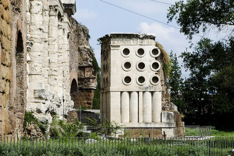 A square monument of pale stone in front of the gate's piers, its upper part pierced by rows of round openings and its lower part formed of close-set vertical cylinders, behind a low railing.