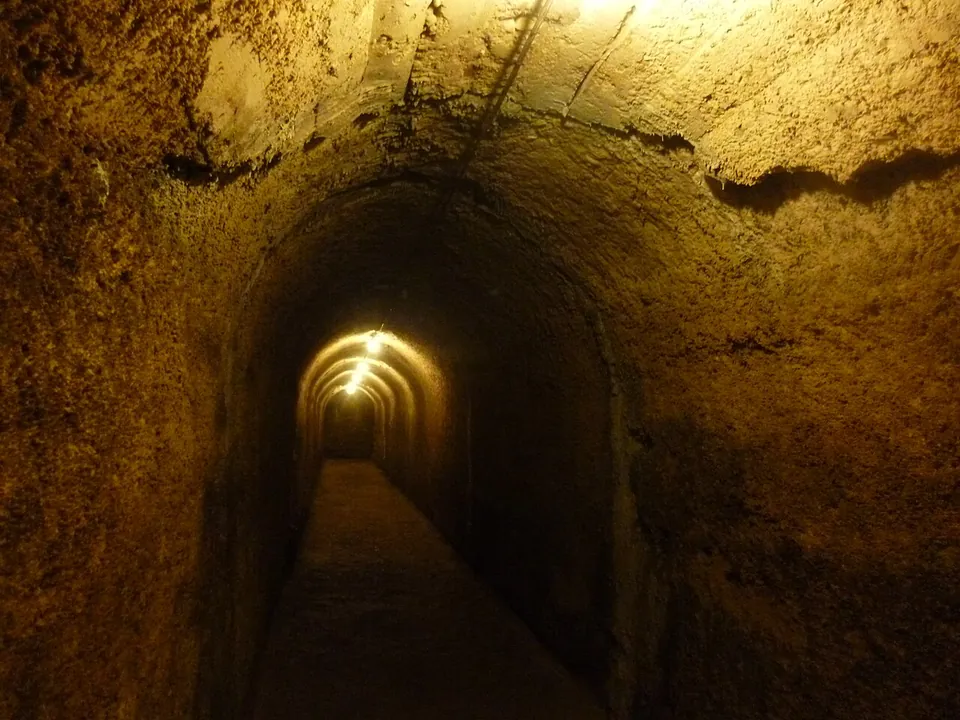 A long barrel-vaulted tunnel with rough plastered walls, lit by a row of lamps receding into the distance.