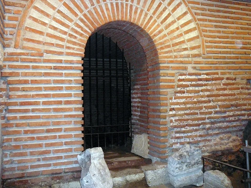A round-headed archway of red brick, its doorway closed by an iron grille, set in a wall of brick and stone, with fragments of carved white stone on the ground in front.