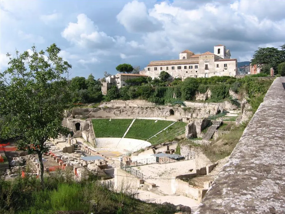 The theatre seen from a stone parapet above, its curved seating banks grassed over and cut by stairways, with the excavated stage building and rows of stone fragments in the foreground and town buildings on the hill behind.