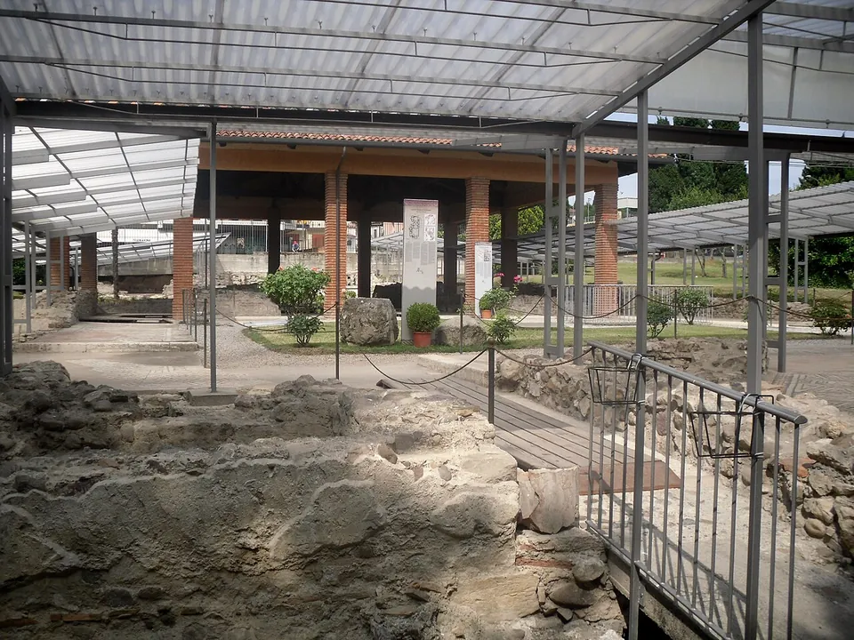 Low ruined walls and floor levels under a long metal-framed canopy, crossed by a wooden walkway with railings, with a row of square brick-red pillars and a lawn beyond.