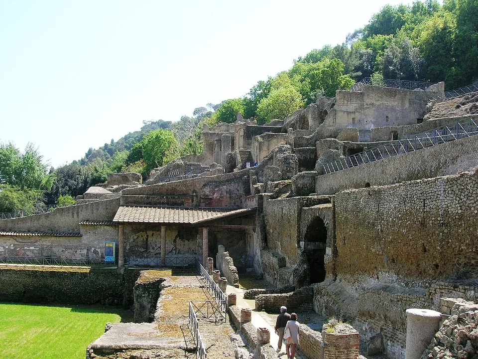 Tall walls of brick and stone stepping up a wooded slope in several levels, with railings along the upper terraces, a tiled shelter and a green lawn below, and two visitors walking on a path at the bottom.