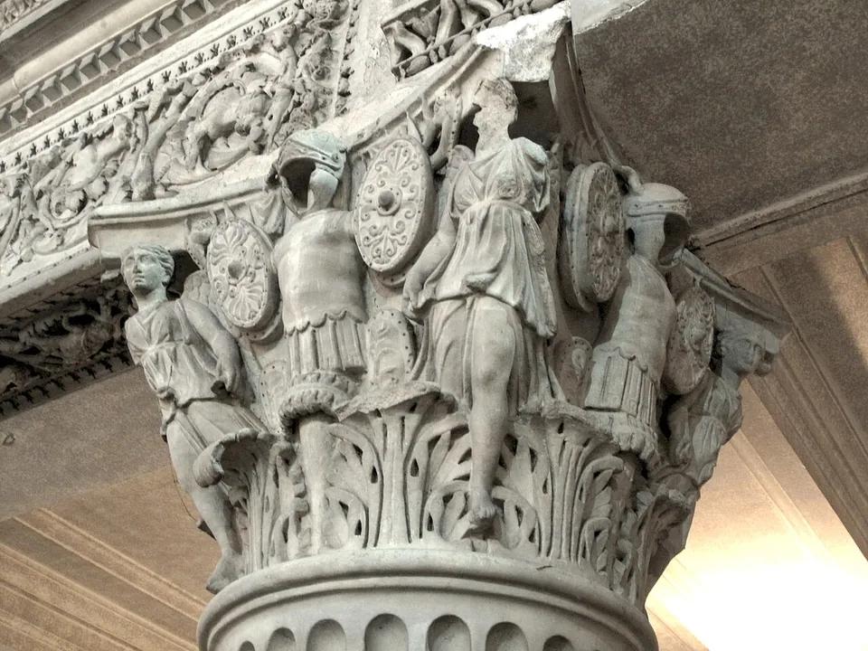 Close view of a marble column capital carved with leaves below and, above them, figures in tunics standing among helmets, round shields and body armour, under a carved beam.
