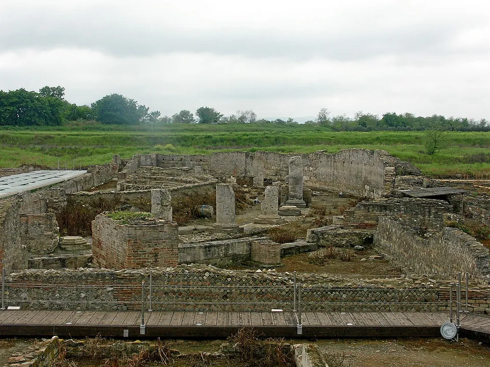 Low ruined walls of rubble and brick and a few square stone piers spread across a sunken excavation, seen from a wooden boardwalk with a railing, with flat green farmland and trees beyond.
