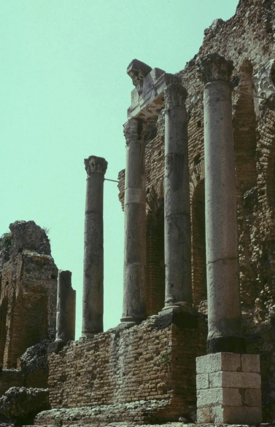 Tall stone columns with carved leafy capitals standing on a brick base in front of a high wall of brick arches, with a fragment of carved entablature resting on two of them.