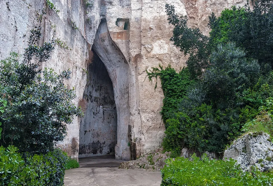A tall, narrow cave mouth tapering to a point at the top, cut into a sheer pale rock face hung with shrubs and creepers, with a paved path leading into the dark inside.