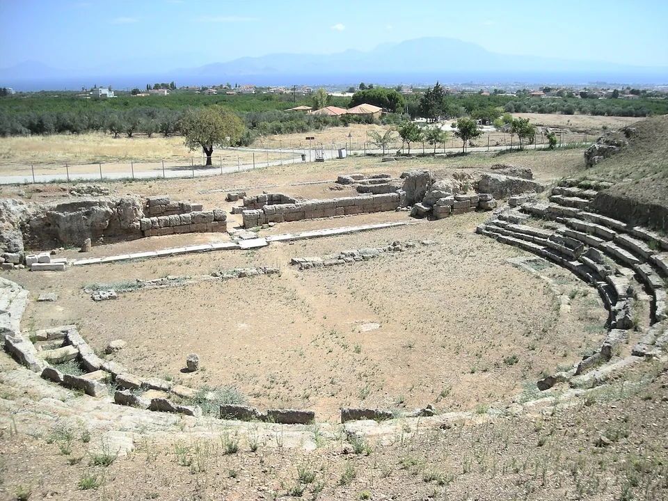 A large theatre hollowed out of a hillside, its lower curved rows of stone seats around a wide earthen orchestra, with the low walls of the stage building beyond, then orchards, the plain and mountains across the sea.