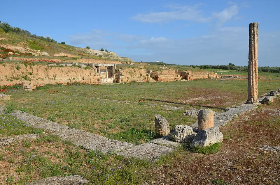 A green field crossed by low stone foundations and a paved line, with a single tall column on the right and an earthen terrace behind holding a small white restored structure with columns.