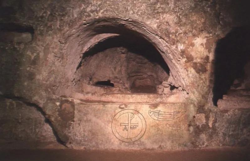 An arched recess cut into the rock above a smoothed wall incised with a circled Chi-Rho monogram between the letters alpha and omega, with a second incised figure beside it.