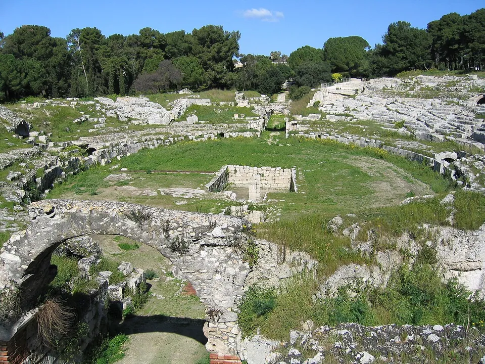 The oval arena of the amphitheatre, its lower tiers cut from pale rock and overgrown with grass, a rectangular stone-lined pit in the middle of the arena and a vaulted passage in the foreground, with pine woods behind.