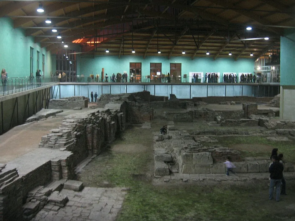 The inside of a long modern hall with pale green walls and a dark beamed roof. Low walls of brick and stone stand on the excavated floor, with visitors along a railed walkway round the edge and a few standing among the ruins below.
