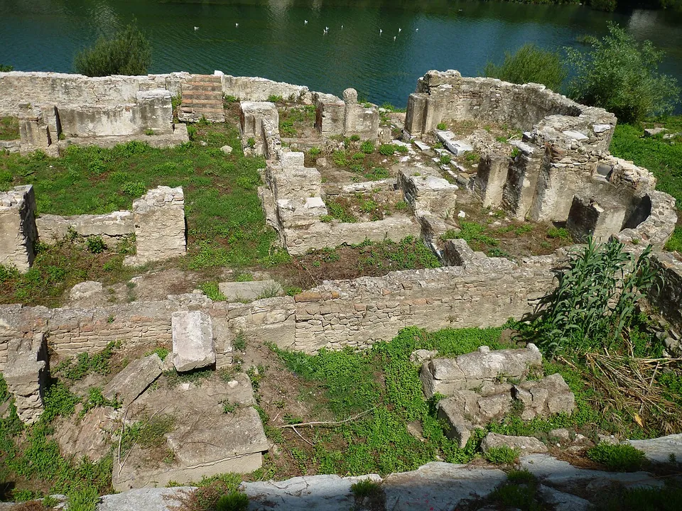 Low walls of squared stone blocks laid out in rooms and a rounded apse among grass and weeds, with the water of a river directly behind them.