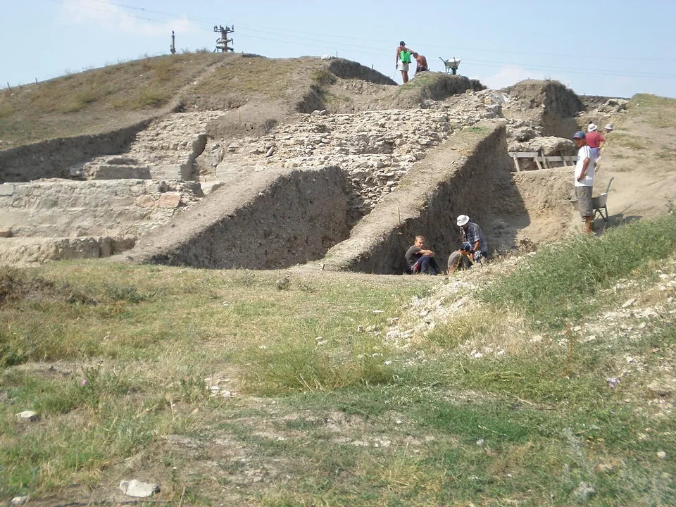 Excavation trenches cut into a grassy slope, with walls of stone and brick exposed in them and archaeologists at work, under a blue sky crossed by power lines.