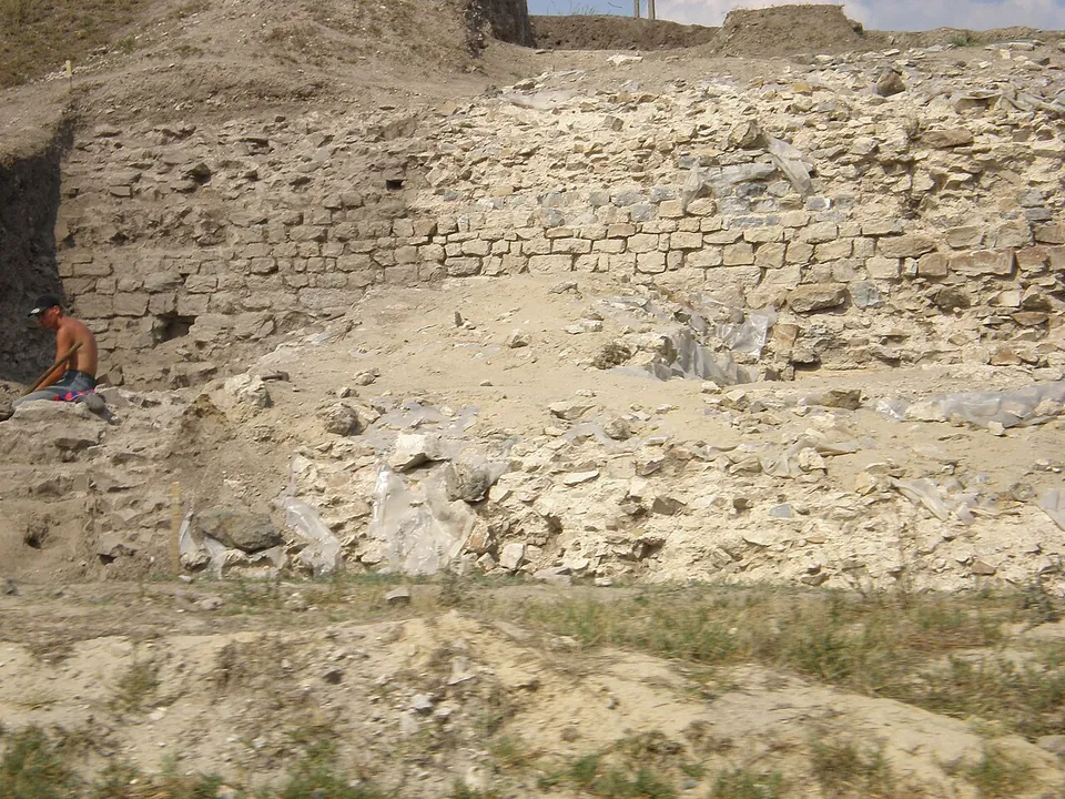 A tall stretch of wall of rough stone with several courses of brick, exposed in an excavation trench, with a man sitting at its foot.