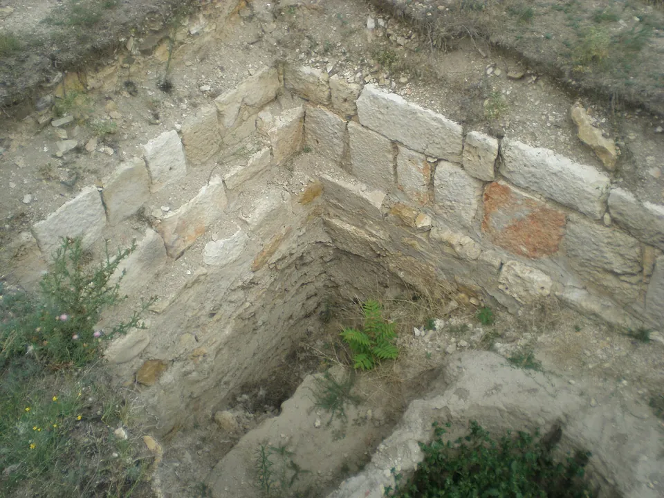 The corner of a sunken room lined with squared pale stone blocks, with a curved stone edge below and weeds growing between the stones.