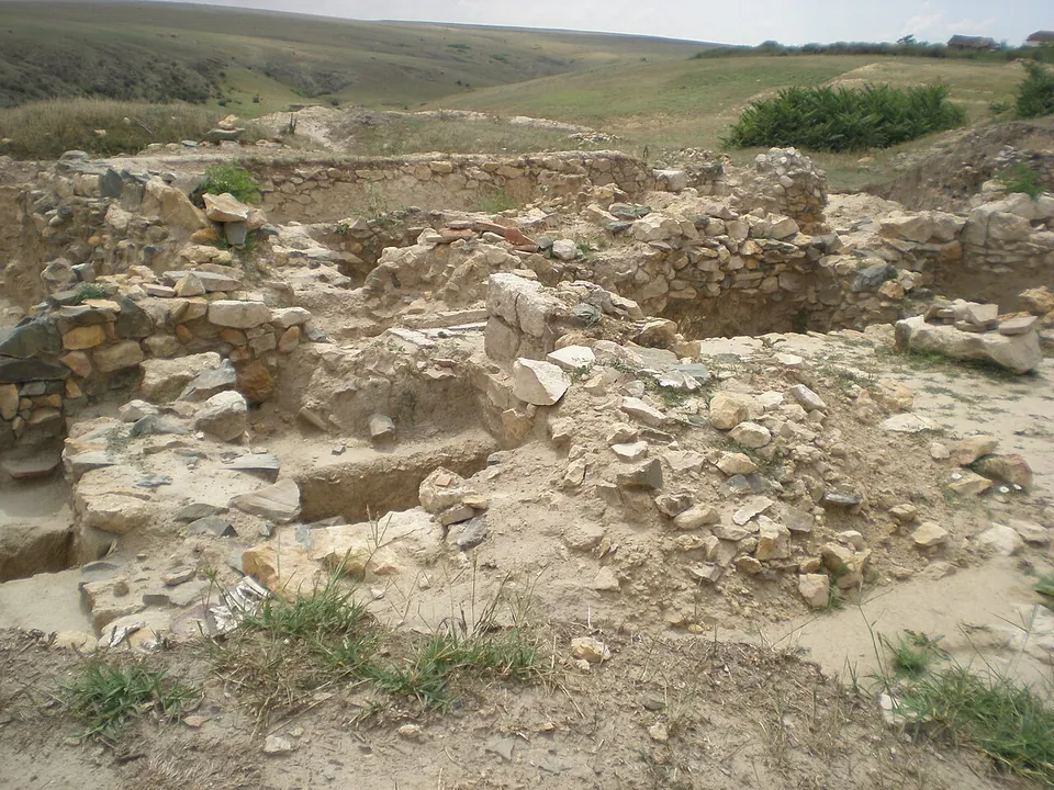 Rubble walls and fallen stones of excavated buildings in the foreground, with a dry grassy plateau and bushes beyond under a pale sky.