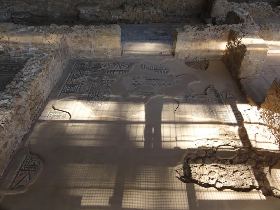 A mosaic floor between low rubble walls, framed by geometric borders with a band of lettering, partly in the shadow of a roof grid.