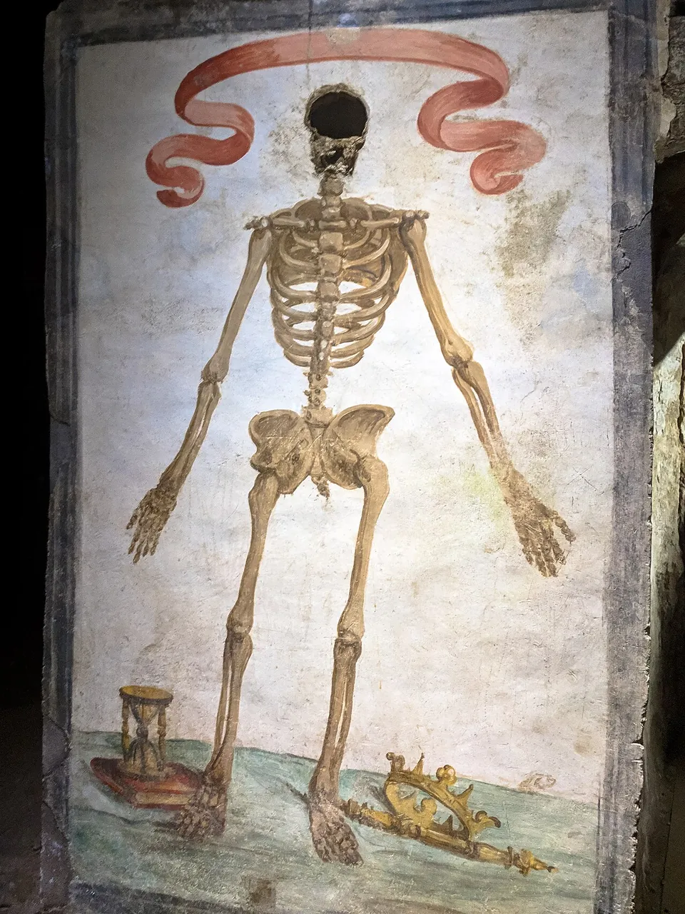 A painted skeleton standing on a plastered wall panel under a red ribbon, with a dark hole where its head should be; an hourglass stands by its left foot and a crown and sceptre lie by its right.