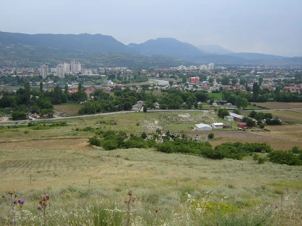 A grassy hillside in the foreground looking down on a fenced excavation area with low walls and a few sheds, the tower blocks and houses of Skopje beyond and mountains on the horizon.