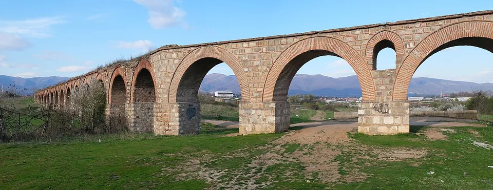A long arcade of round brick arches on massive stone piers crossing a grassy field, with a dirt track passing under one arch and mountains on the horizon.