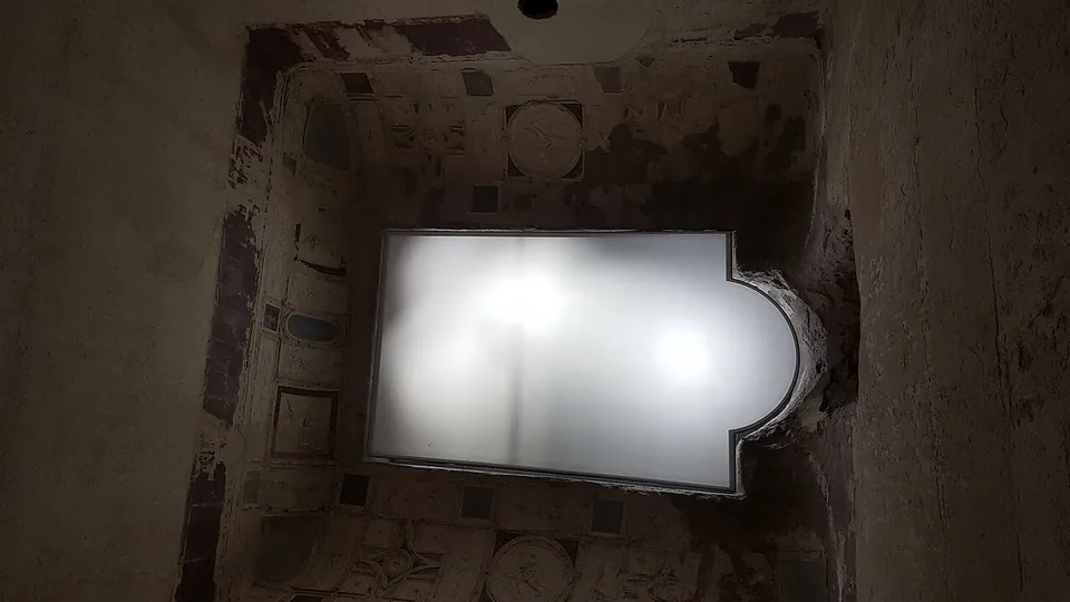 Looking up into the vestibule's vault, an opening in the ceiling glazed with white frosted glass, framed by stucco panels and roundels on a ground of dark red and green-grey paint.